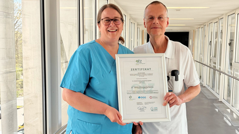 Dr. Saskia Otte, Chefärztin der Geriatrie und Physikalischen Medizin, und Dr. Olaf Pingen, Chefarzt der Orthopädie und Unfallchirurgie, stehen im Amalie Sieveking Krankenhaus in Hamburg-Volksdorf in einem hellen Durchgang und halten gemeinsam das Zertifikat für die erfolgreiche Zertifizierung des Krankenhauses als Alterstraumatologisches Zentrum in die Kamera. Dr. Saskia Otte, Chefärztin der Geriatrie und Physikalischen Medizin, und Dr. Olaf Pingen, Chefarzt der Orthopädie und Unfallchirurgie, stehen im Amalie Sieveking Krankenhaus in Hamburg-Volksdorf in einem hellen Durchgang und halten gemeinsam das Zertifikat für die erfolgreiche Zertifizierung des Krankenhauses als Alterstraumatologisches Zentrum in die Kamera.